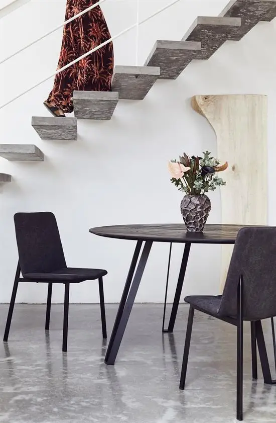 Moderne spisestue med rundt bord, sorte stole, betontrappe og vase med blomster.