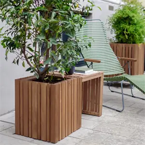 Træbord med plante i kvadratisk kasse, grøn stol og ekstra plantekasse i baggrunden.