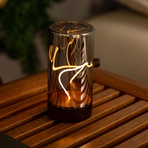 Cylindrisk bordlampe i reflekterende metal med dekorativ lysfilament på træoverflade.