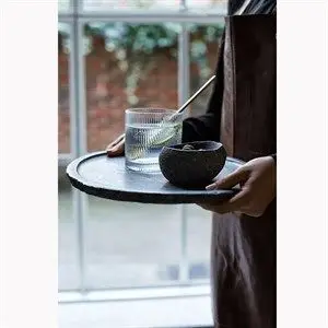 Person holder bakke med ribbet Muubs-glas og skål. Glas med drik og grønt. Sløret vinduesbaggrund.