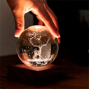 Hånd rører krystalkuglelampe med 3D hjort, lyser varmt på træbase.