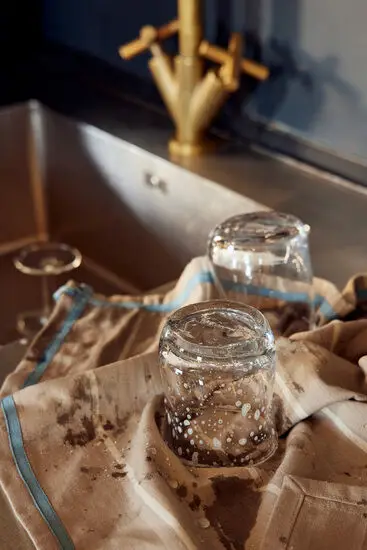 Beige og blåstribet viskestykke med glas ved vask. Hængestrop og broderet logo ses.