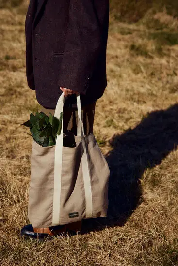 Beige totebag med lange hanke, bæres af person, indeholder grønne blade.