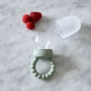 Babyfrugtfeeder med grøn hank, gennemsigtig pose, låg ved siden af og tre hindbær på overfladen.