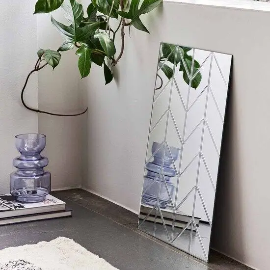 Rektangulært spejl med sildebensmønster, lænet mod væg. Plante og dekorativ vase ved siden af.