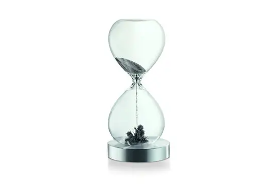 Timeglasformet objekt med klar krop, mørkt granulat indeni, på en cirkulær metalbase.