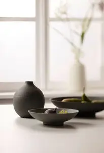 Rund vase med horisontale riller, stående ved skåle med frugt i minimalistisk indretning.