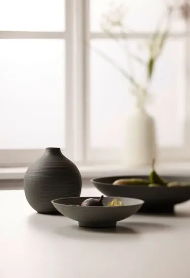 Rund vase med horisontale riller, stående ved skåle med frugt i minimalistisk indretning.