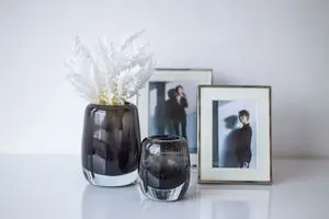 Philippi "Emma" vase S, sort og skinnende, ved siden af større vase med hvide blomster og to indrammede fotos.
