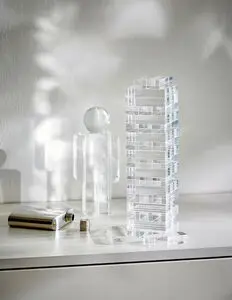 Klar akryltårn med rektangulære blokke, figur ved siden af, og metallisk flaske med låg på bordet.