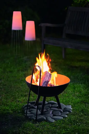 Rund ildskål på ben, omgivet af sten. Flammer lyser op. Bænk og lamper i baggrunden.