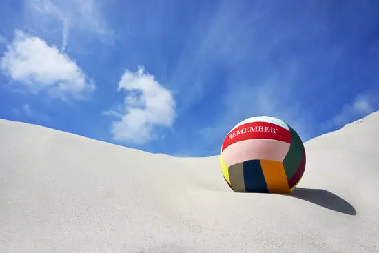 Bold med farverige segmenter, ordet "REMEMBER", ligger på sand mod blå himmel.
