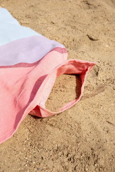 Farverigt strandhåndklæde på sand med stofløkke i hjørnet.