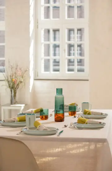 Spisestue med lys borddækning, lime bomuldsservietter på tallerkener, glas og bestik i matchende stil.