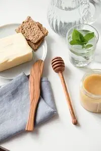 Smørkniv og honningske i oliventræ på bord med smør, knækbrød, honning og myntevand.