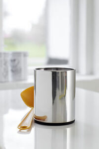 Skinnende stålredskabsholder med sort bund og gul spatula på blank overflade.