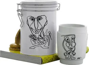 Hvid beholder og krus med sort kunstnerisk ansigtstegning, placeret ved siden af hinanden.